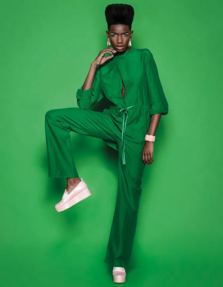 A model in a green jumpsuit against a flat green backdrop, one knee raised, in pale pink platform shoes