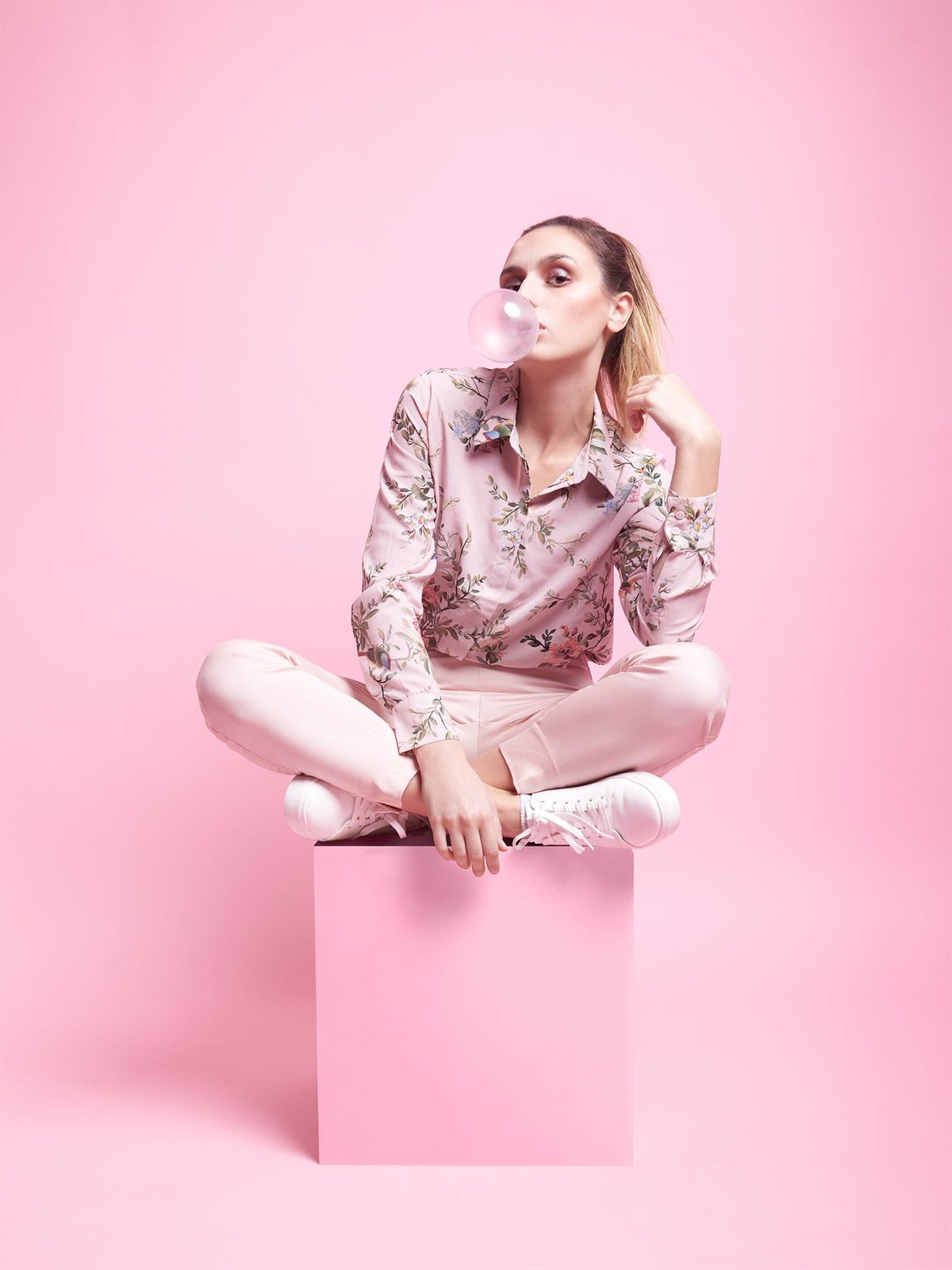 Model in a pink floral blouse and matching trousers blowing a bubblegum bubble, seated cross-legged on a pink pedestal against a pink background