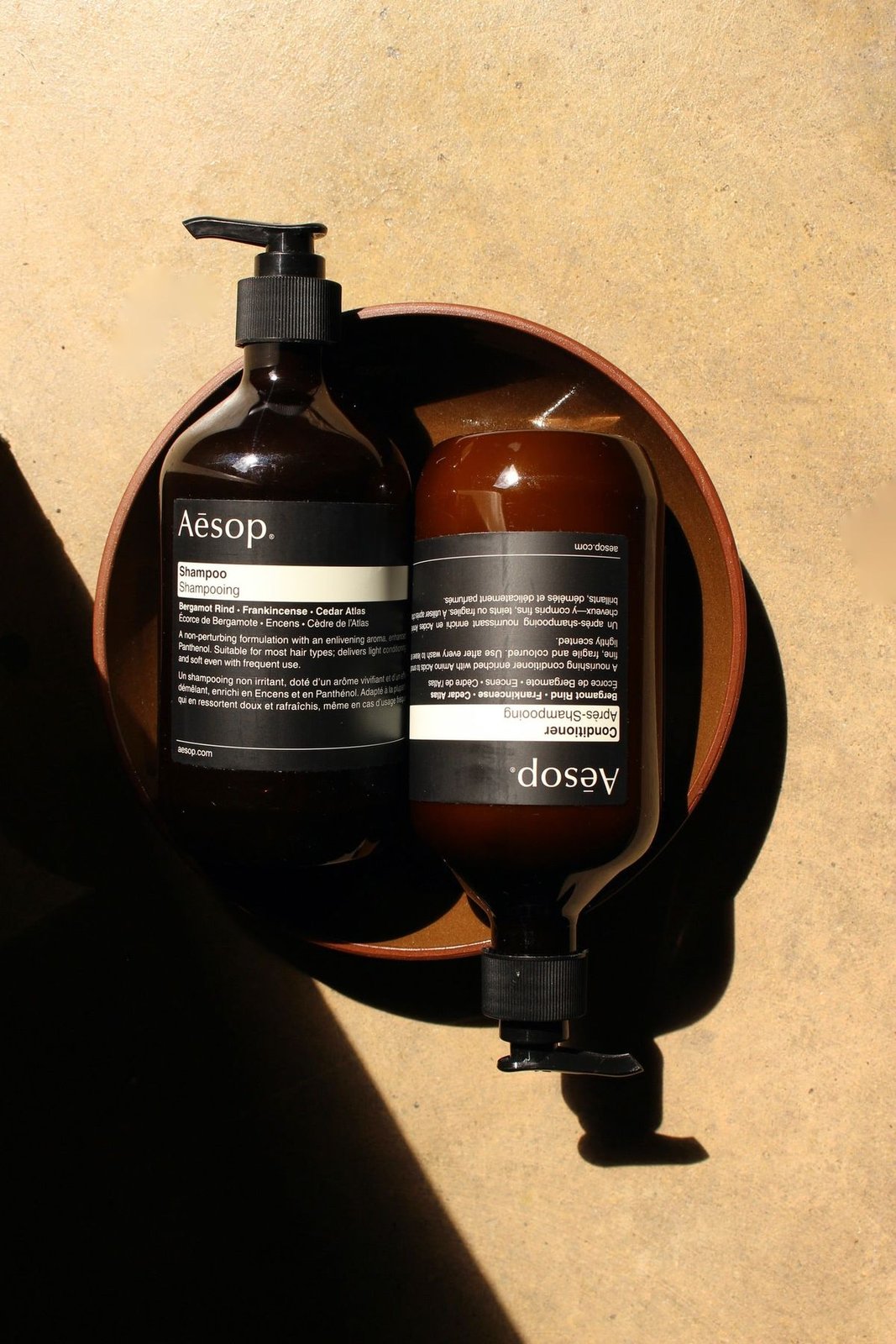 Aesop shampoo and conditioner bottles resting in a round leather tray, lit by warm directional sunlight with strong shadow